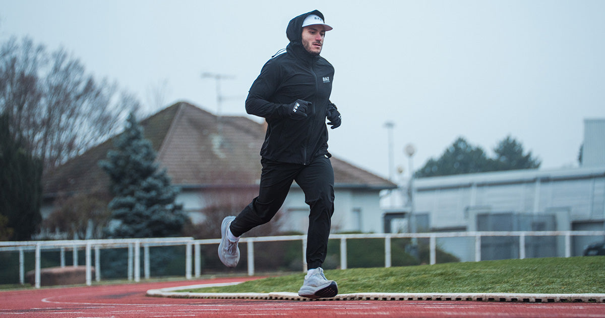 Athlète homme en jogging et coupe-vent Baz Athletics en train de courir sur piste  tenue de running homme performance